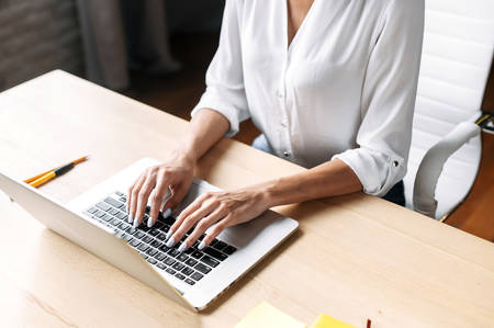 Female hands on the keyboard of the trendy laptop indoor, a dark skinned woman wearing white formal shirt, a face is not visible. Close-up photographyの写真素材