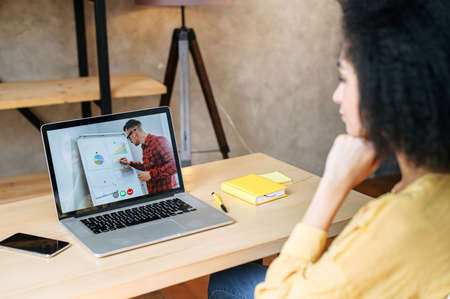 Webinars, lectures online. An african american woman in office is watching online classes, male teacher with a flip chart on a laptop screen. Professional development, business trainingの写真素材