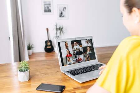 Video call. A young woman communicates via video. She is sitting at the table in cozy living room, looks at laptop screen. Back viewの写真素材