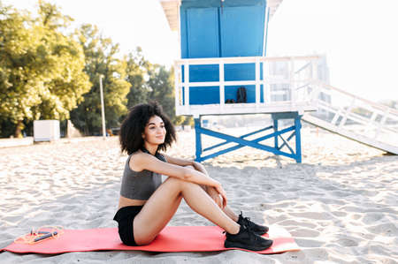 A beautiful biracial girl is resting after workout sitting on the mat at the beach. A young mixed-race woman in sportswear sits and looks awayの写真素材