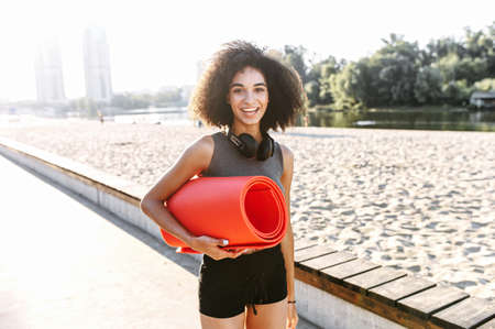 A smiling mixed-race young woman with a rolled gymnastics mat in hands on the city waterfront. Sporty lifestyle conceptの写真素材
