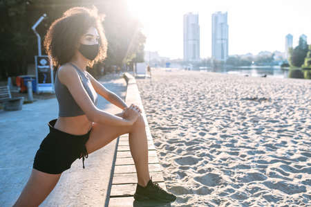 A beautiful athletic African-American young woman with a medical mask on the face is warming up before morning jogging outdoor the city waterfront. Sports during quarantineの写真素材