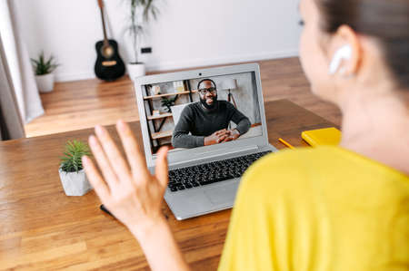 Video call. A young woman sits in the living room and uses laptop for video connection with a friend, coworker, she talks with an african-american guyの写真素材