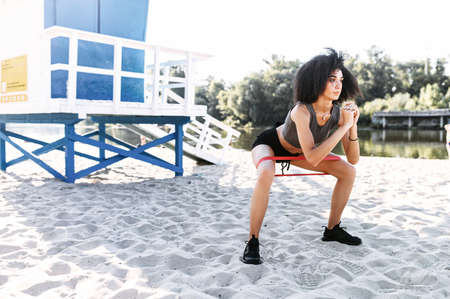 An attractive athletic mixed-race woman doing outdoors workout with a resistance rubber band. A girl in sportswear is training on the beachの写真素材