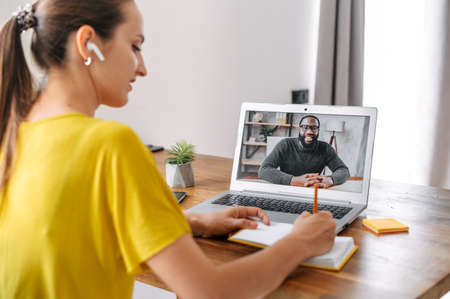 Video call. A young woman sits in the living room and uses laptop for video connection with a friend, coworker. Woman writes notes and talks with an african-american guyの写真素材