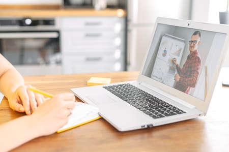 Female hands is writing in notebook while male teacher, tutor, coach on the laptop conducts online training, webinar. Online learning conceptの写真素材