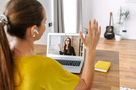 A young woman sits indoor and using app on laptop for video call to female coworker, colleague or friend. Two young women in casual wear talking online via video connectionの写真素材