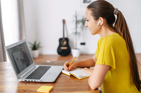 Webinars, online learning, online classes. A female student using laptop for watching lectures online, a woman sits at the table and writing in notebook, a male teacher with flip chart on the screenの写真素材