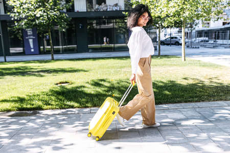 A biracial young woman is walking outdoor with trolley bag looking at the camera with a smile. Side viewの写真素材