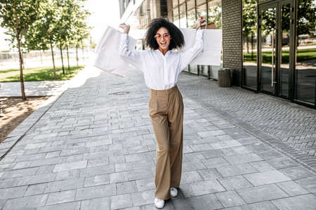 A cheerful mixed-race young woman holds hands up with a many paper shopping bags, a girl with an Afro hairstyle happy with purchases. Full lengthの写真素材