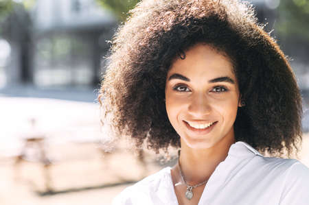 Close-up portrait of curly dark skinned young woman in white shirt, she looks at camera with a smileの写真素材