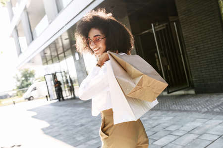Attractive young African-American woman walks on a shopping spree in the city. Beautiful trendy girl carries paper shopping bags and looks back with a smileの写真素材