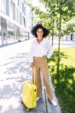 A trendy multi-ethnic business woman with a trolley bag outdoors. A full length shot of girl with a yellow suitcase outdoorsの写真素材