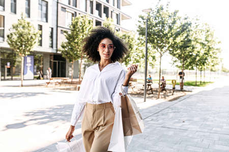 Trendy young African-American woman is carrying shopping bags on the city street. A midshot of multi ethnic girl after good shopping day outdoorの写真素材
