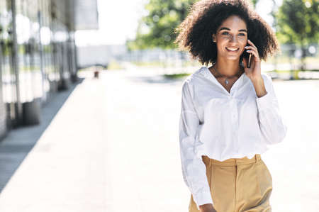 Busy mixed-race woman is talking on the smartphone outdoors. A girl with an Afro hair is walking outdoors and speaks on the mobile phoneの写真素材