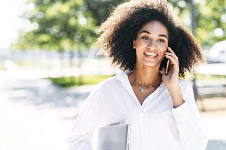 Confident African-American woman in white shirt with a laptop in hand talking on the smartphone outdoorsの写真素材