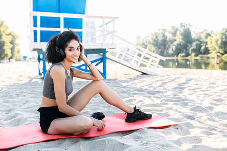 An athletic beautiful biracial girl in sportswear listens music and rests on the beach. A woman with an afro hair sits on the gymnastic mat outdoorの写真素材