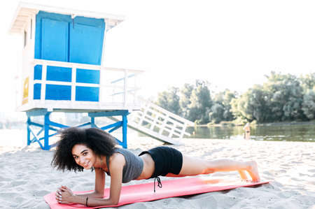 An African-American girl in sportswear is training outdoors. A woman is doing plank exercise on the gymnastic mat on the beachの写真素材