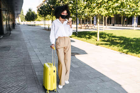 An African-American businesswoman wearing medical mask with a suitcase check in flight on a smartphone. Business travel during pandemicの写真素材