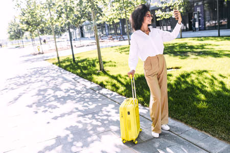An African-American young woman takes a selfie before journey. A girl with an Afro hairstyle with a yellow suitcase outdoor using smartphoneの写真素材