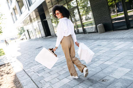 Attractive young African-American woman walks on a shopping spree in the city. Beautiful trendy girl carries paper shopping bags and looks back with a smile. Full lengthの写真素材