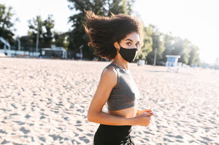 Midshot of a young multi-ethnic woman with a medical mask is running outdoors. Morning exercises during pandemic, quarantineの写真素材