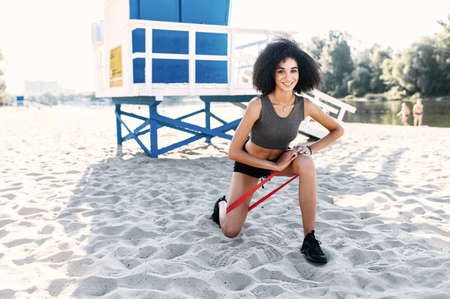 A slender fresh African girl is doing lower body workout with a resistance rubber band outdoor on the city beach, a rescue point on the backgroundの写真素材