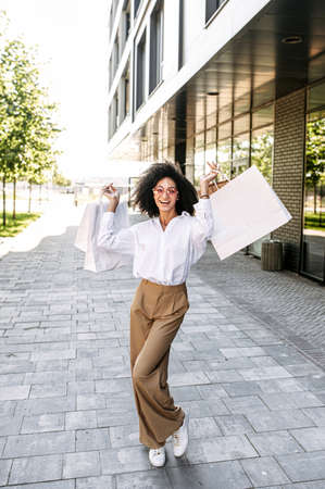 A cheerful mixed-race young woman holds hands up with a many paper shopping bags, a girl with an Afro hairstyle happy with purchases. Vertical shotの写真素材
