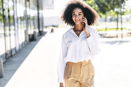 Busy mixed-race woman is talking on the smartphone outdoors. A girl with an Afro hair is walking outdoors and speaks on the mobile phoneの写真素材