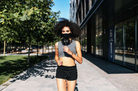 Front view of an African-American girl wearing a protective mask on the face with an afro hairstyle is jogging on the city street. Morning jog during quarantine, pandemicの写真素材