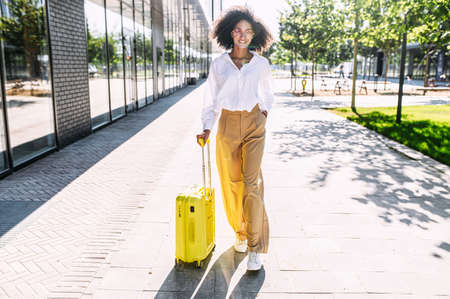 An African-American young woman going to travel. A girl with an Afro hairstyle with a yellow suitcase walks in the cityの写真素材