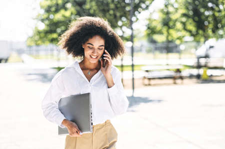 Beautiful african woman is talking on a mobile phone while going to the office with a laptop. Young freelancer girl is calling to the clientsの写真素材