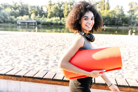 Side view of a smiling African-American girl with a rolled gymnastics mat in hands, she is walking and looks at camera outdoors.の写真素材