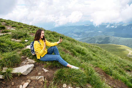 Cheerful girl traveler in communicates by video call on smartphone on top of mountain. Female hiker takes a selfie on mobile phone against background of scenic natureの写真素材