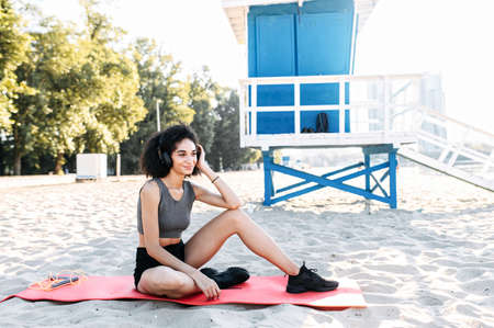 An athletic beautiful biracial girl in sportswear listens music and rests on the beach. A woman with an afro hair sits on the gymnastic mat outdoorの写真素材