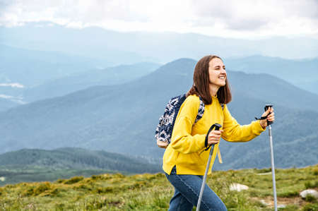 A cheerful hiker girl in yellow sweater with a backpack walks with a sticks on mountains footpath with a scenic view on the backgroundの写真素材