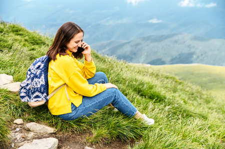 A hiker woman is talking on the smartphone while sitting on the top of the mountain. Backpacker girl speaks on the cellphone and smilesの写真素材