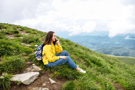 A hiker woman is talking on the smartphone while sitting on the top of the mountain. Backpacker girl speaks on the cellphone and smilesの写真素材