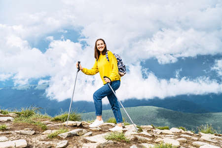 A cheerful hiker girl in yellow sweater with a backpack walks with a sticks on mountains footpath with a scenic sky view on the backgroundの写真素材