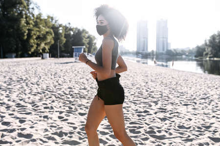 A young mixed-race woman with a medical mask is jogging outdoors. Morning exercises during pandemic, quarantine. Side viewの写真素材
