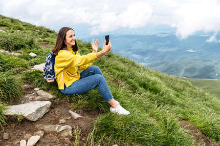 Cheerful girl traveler in communicates by video call on smartphone on top of mountain. Female hiker takes a selfie on mobile phone against background of scenic natureの写真素材