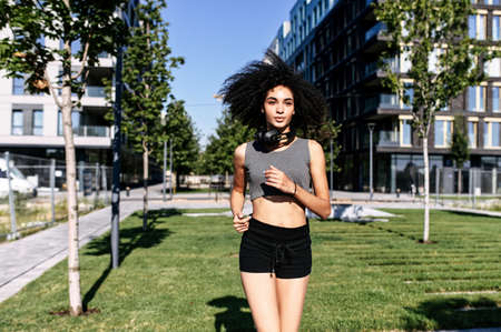 An athletic African-American young woman is jogging outdoors. A girl with an afro hairstyle in short sports bra and shorts runs on the street. Healthy lifestyle concept. Front viewの写真素材