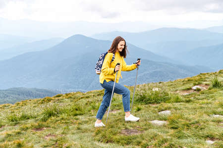 A cheerful hiker girl in yellow sweater with a backpack walks with a sticks on mountains footpath with a scenic view on the backgroundの写真素材