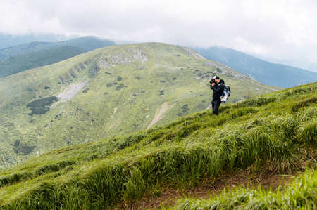 Photographer is taking a photo in the mountains. A hiker guy with a backpack stands on footpath with a professional camera with a scenic view on the backgroundの写真素材