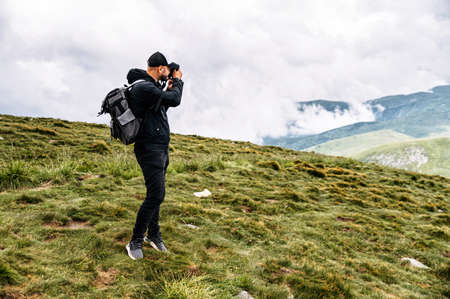 Photographer is taking a photo in the mountains. A hiker guy with a backpack stands on footpath with a professional camera with a scenic view on the backgroundの写真素材