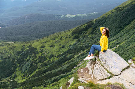 A cheerful woman sits on the top of the cliff and looks at camera with a smile. Scenic view on the mountains on the background. Resting and recreation in the natureの写真素材