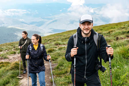 Group of young cheerful people with nordic walking poles on the top of the mountain. A hipster bearded guy in a cap looks at cameraの写真素材