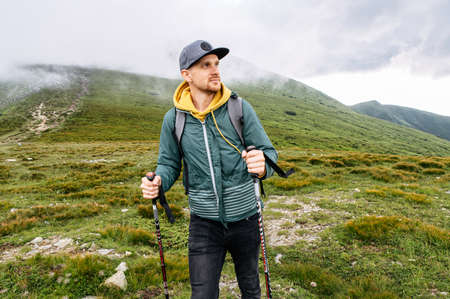 Active backpacker guy engaged nordic walking outdoors. Hipster in cap walks on the footpath on the green hill. Healthy lifestyleの写真素材