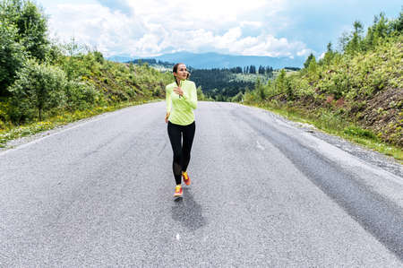 An athletic girl in is running on the road in the mountains. A young woman in trendy sportswear jogging outdoors with a scenic view on the backgroundの写真素材