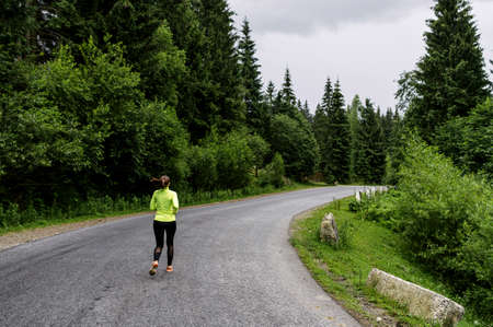 A young woman running outdoors. A girl in trendy long sleeve t-shirt is jogging on the road surrounded pines forestの写真素材